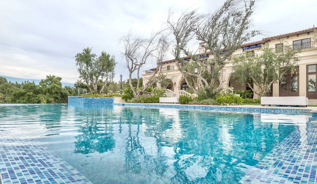 Garden pool with purple flowers, a white bench under a large tree, and mountains beyond